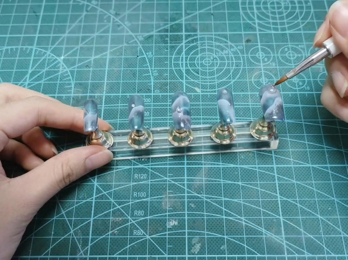 Un dotting tool plaçant une minuscule perle argentée près de la zone cuticulaire dun ongle sur un présentoir.