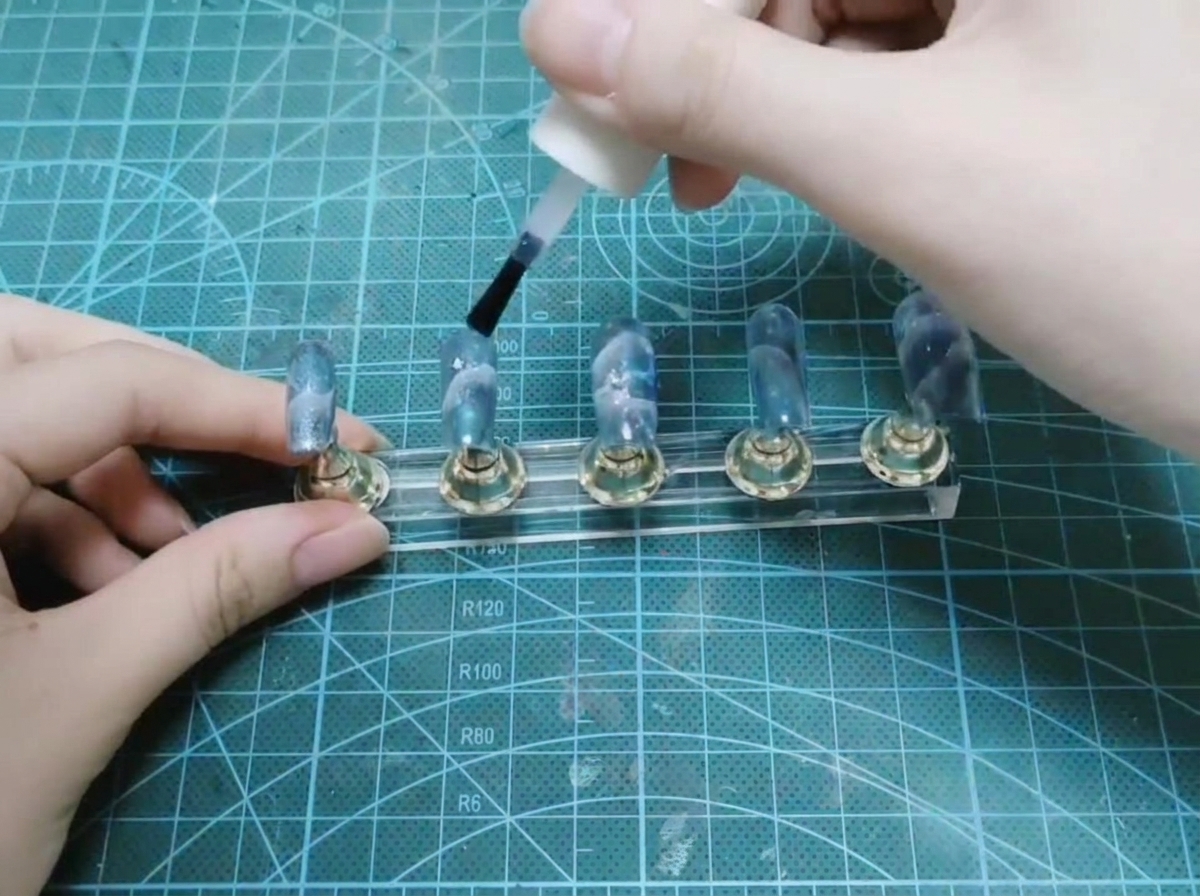 Une main appliquant une finition brillante avec le pinceau dun flacon blanc sur un ensemble dongles terminés sur un présentoir.