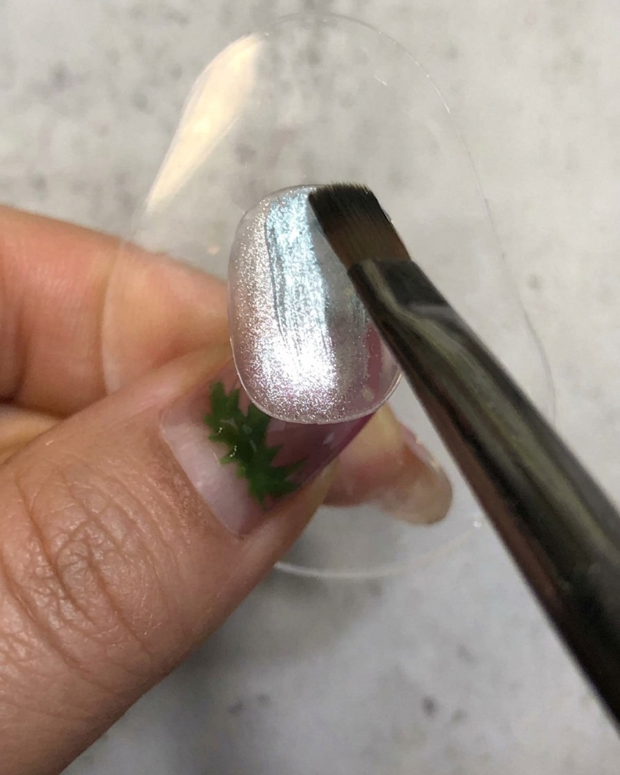 Un pinceau à ongles appliquant un vernis gel bleu diaphane sur une base magnétique argentée brillante.