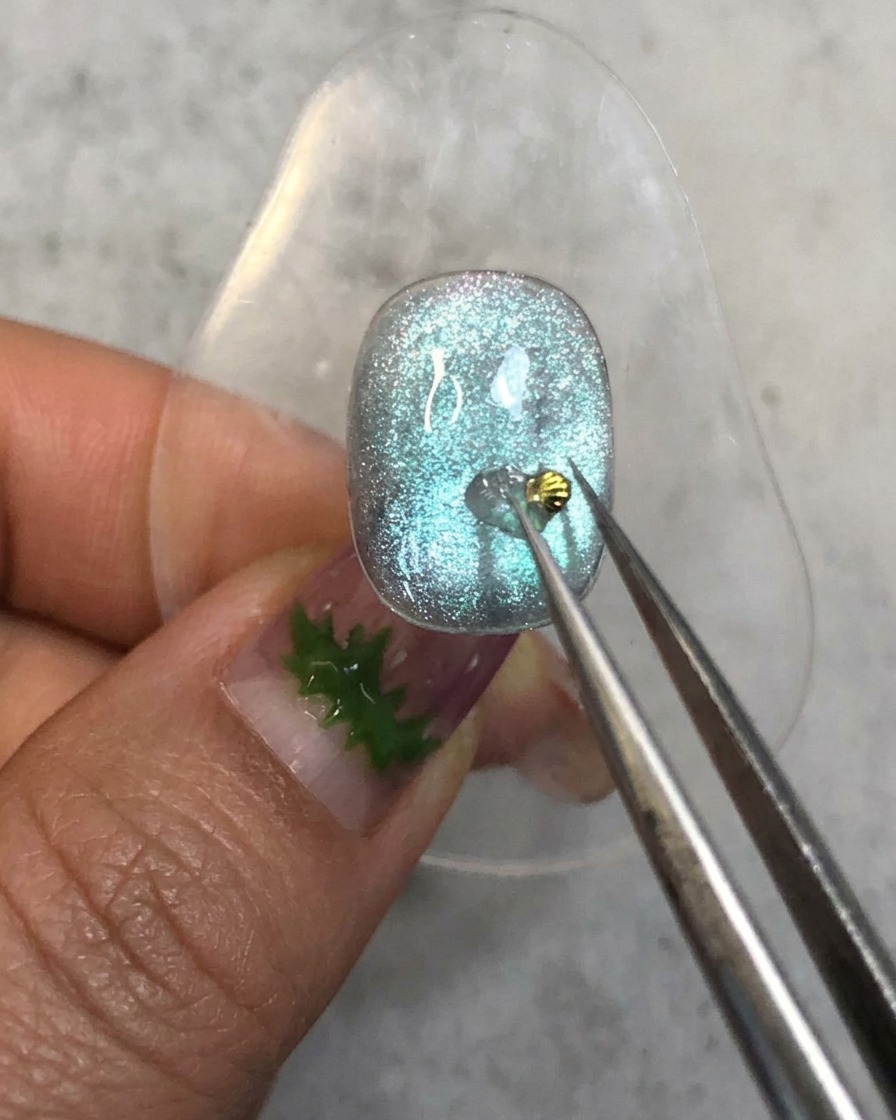 Une paire de brucelles plaquant une minuscule breloque de coquillage doré sur la goutte de gel de renfort sur un ongle bleu.