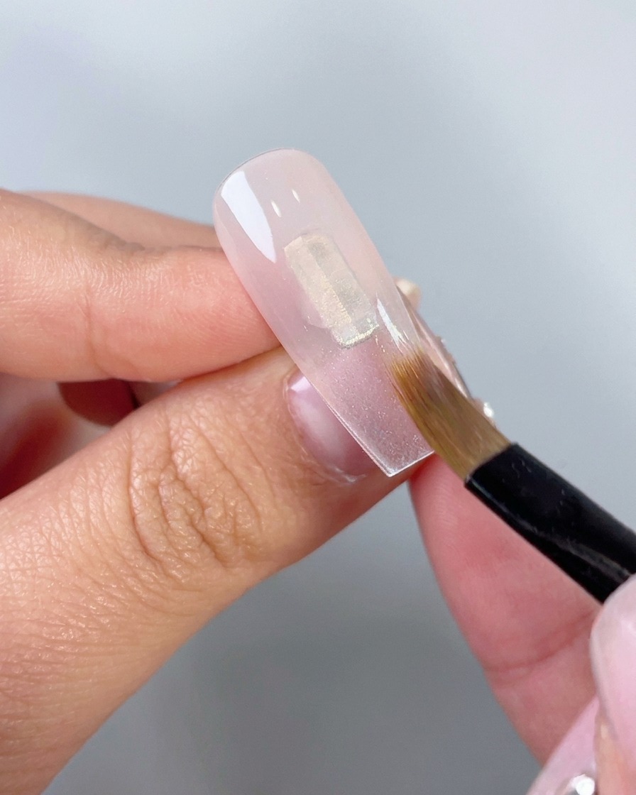 Gros plan dun pinceau de maquillage plat estompant du gel rose translucide vers le bas sur une capsule dongle artificielle transparente.