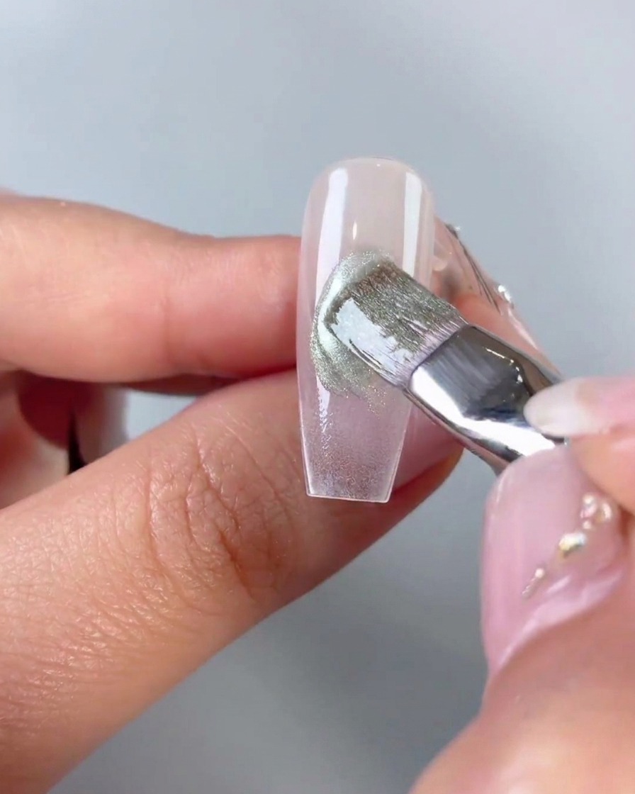 Un pinceau plat lissant et estompant du gel œil-de-chat vert appliqué en diagonale au centre dune capsule dongle transparente.