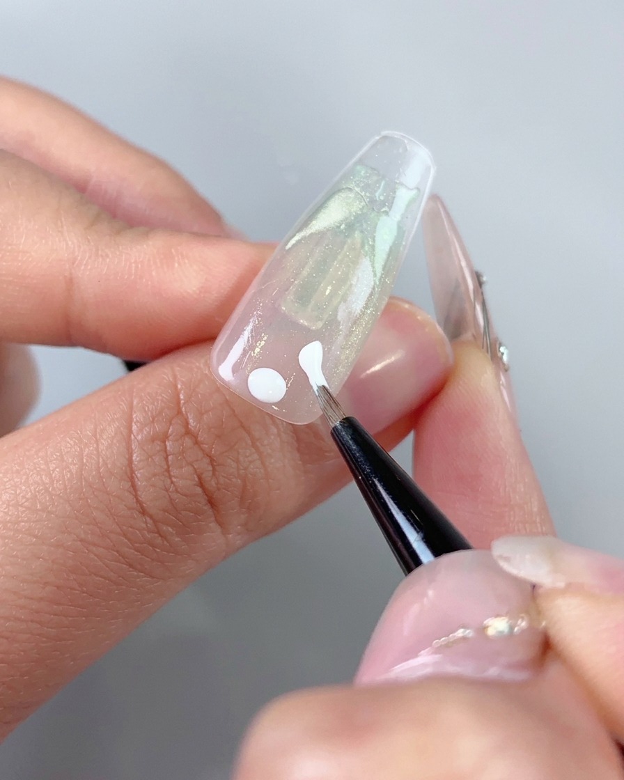 Un pinceau fin appliquant des points de gel blanc et des traits initiaux près de la zone des cuticules dun ongle.