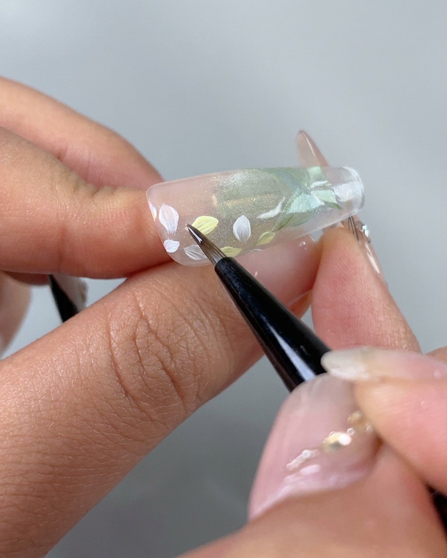 Un pinceau fin ajoutant avec précaution des teintes de gel rose clair et jaune sur des pétales de fleurs blancs peints individuels sur un ongle.