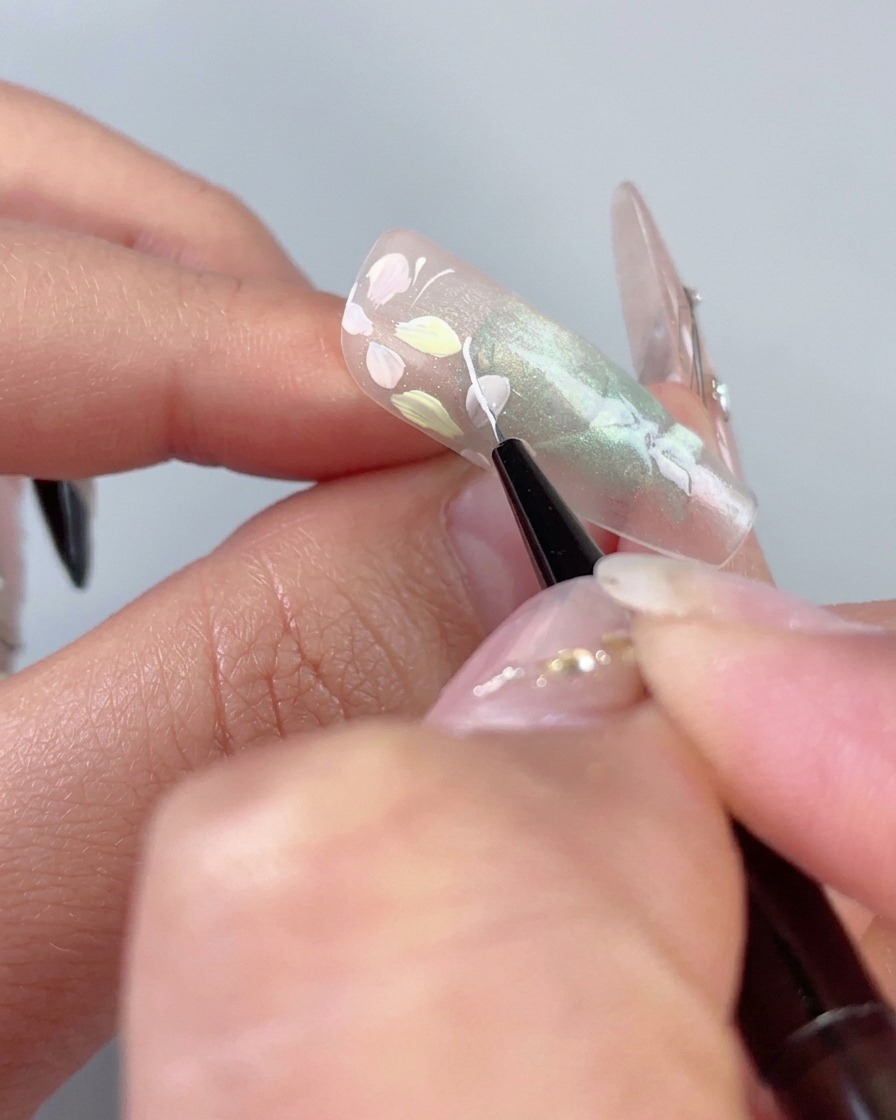 Un pinceau traceur fin dessinant une délicate ligne blanche incurvée à côté de fleurs pastel sur un ongle œil-de-chat vert transparent.