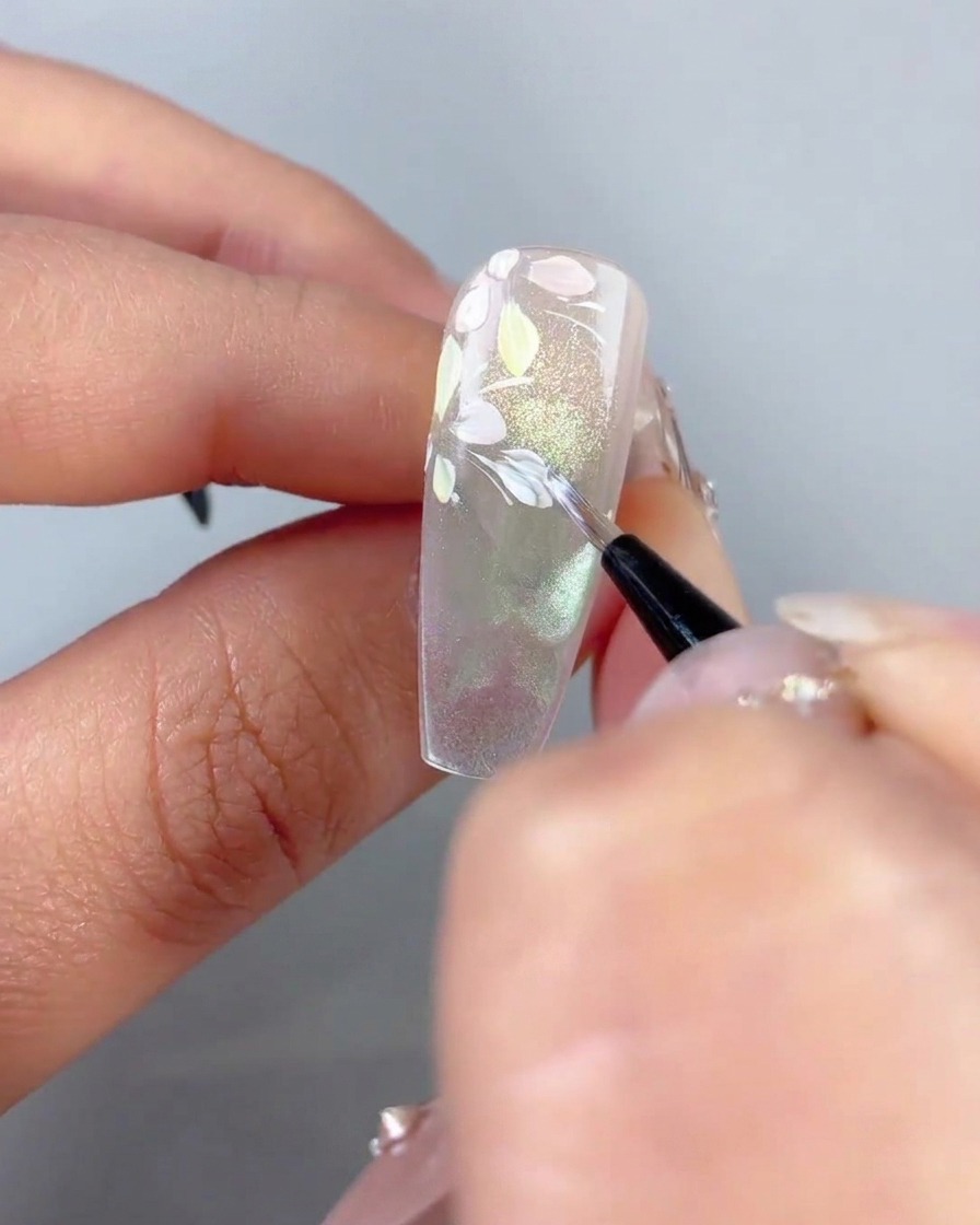 Un pinceau fin déposant de minuscules accents de feuilles blanches sur des lignes blanches incurvées près de pétales de fleurs pastel sur un ongle.