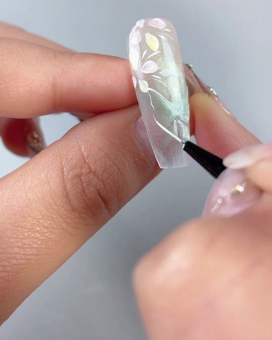 Un pinceau traceur fin traçant de fines contours blancs et des nervures dailes sur une forme de papillon œil-de-chat vert diaphane sur un ongle.