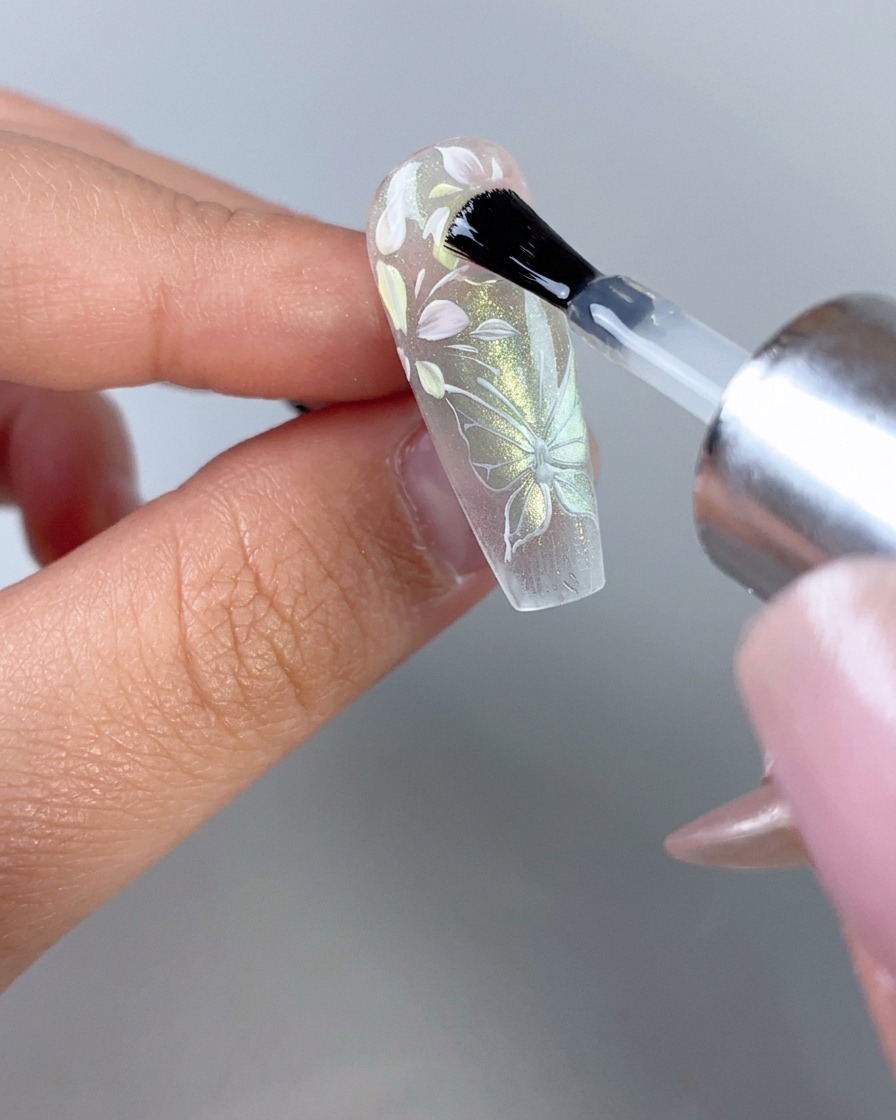 Un pinceau à vernis à ongles appliquant une couche de finition transparente brillante sur un ongle décoré de fleurs et de papillons transparents.