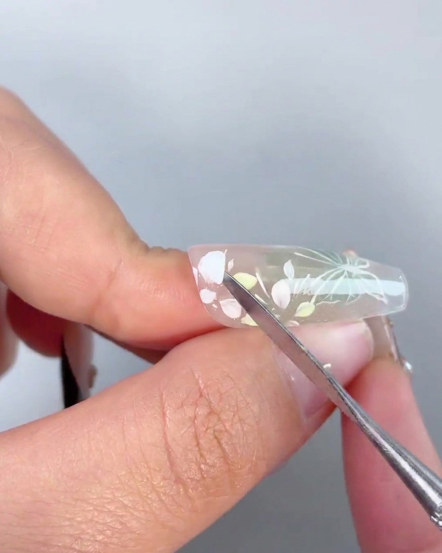 Un outil métallique appliquant une goutte de gel adhésif transparent au centre dun design floral peint sur un ongle.