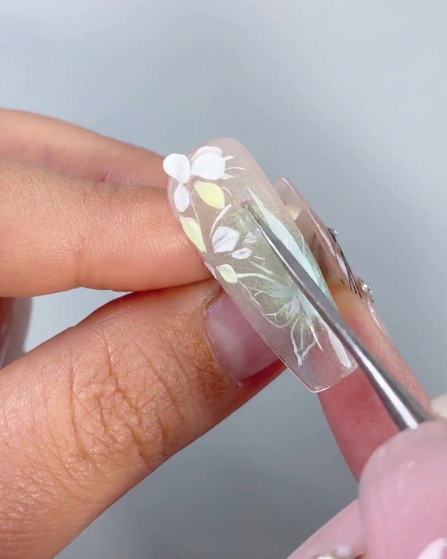 Un outil métallique plaçant un pétale 3D blanc préformé sur le gel adhésif dun ongle.