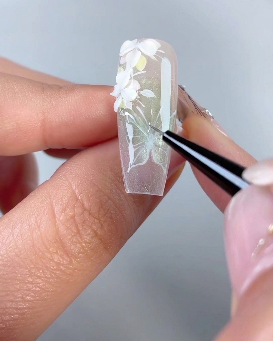 Gros plan dun pinceau traceur fin appliquant du gel de modelage 3D transparent sur les lignes peintes en blanc dun design daile de papillon sur une capsule dongle en gel.