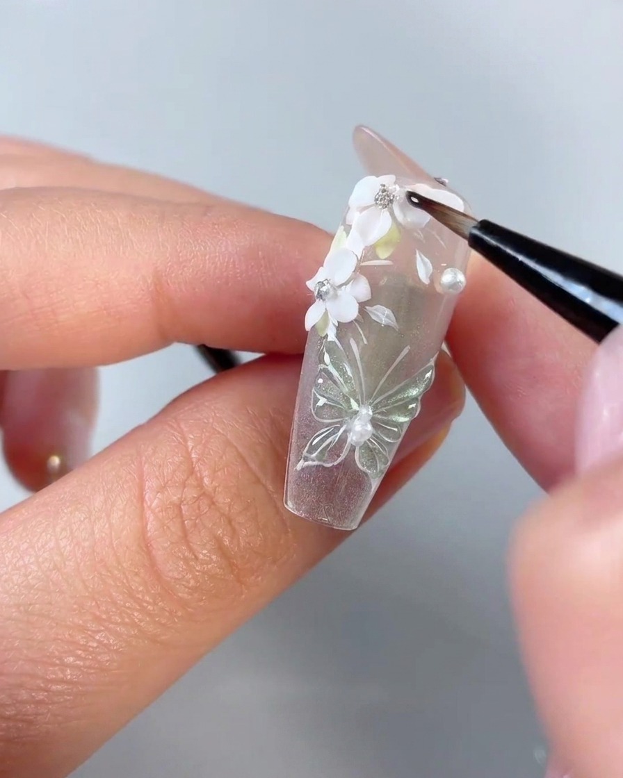 Un pinceau à ongle de précision appliquant du gel transparent autour des bords des pétales de fleurs 3D blancs et des strass sur une capsule dongle transparente.