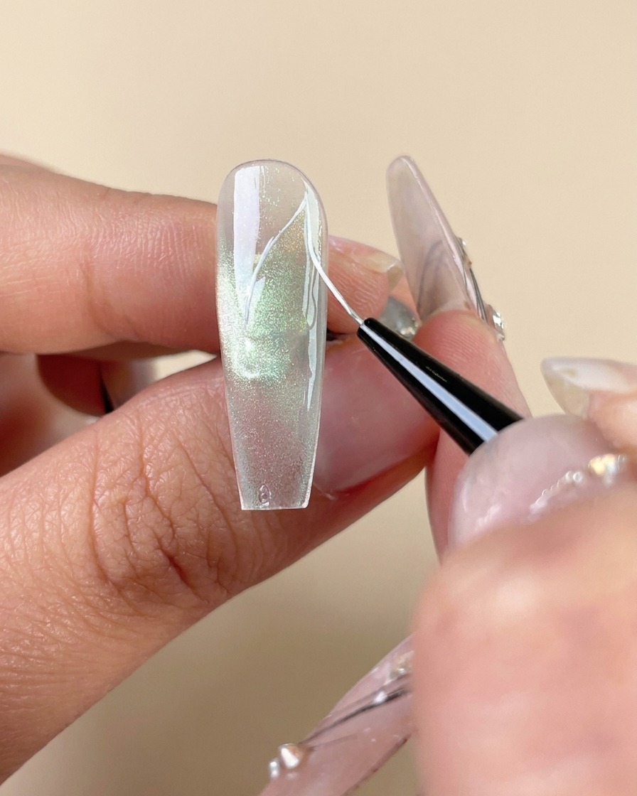 Un pinceau traceur fin dessinant des lignes de nervures blanches sur un ongle en gel œil-de-chat vert.