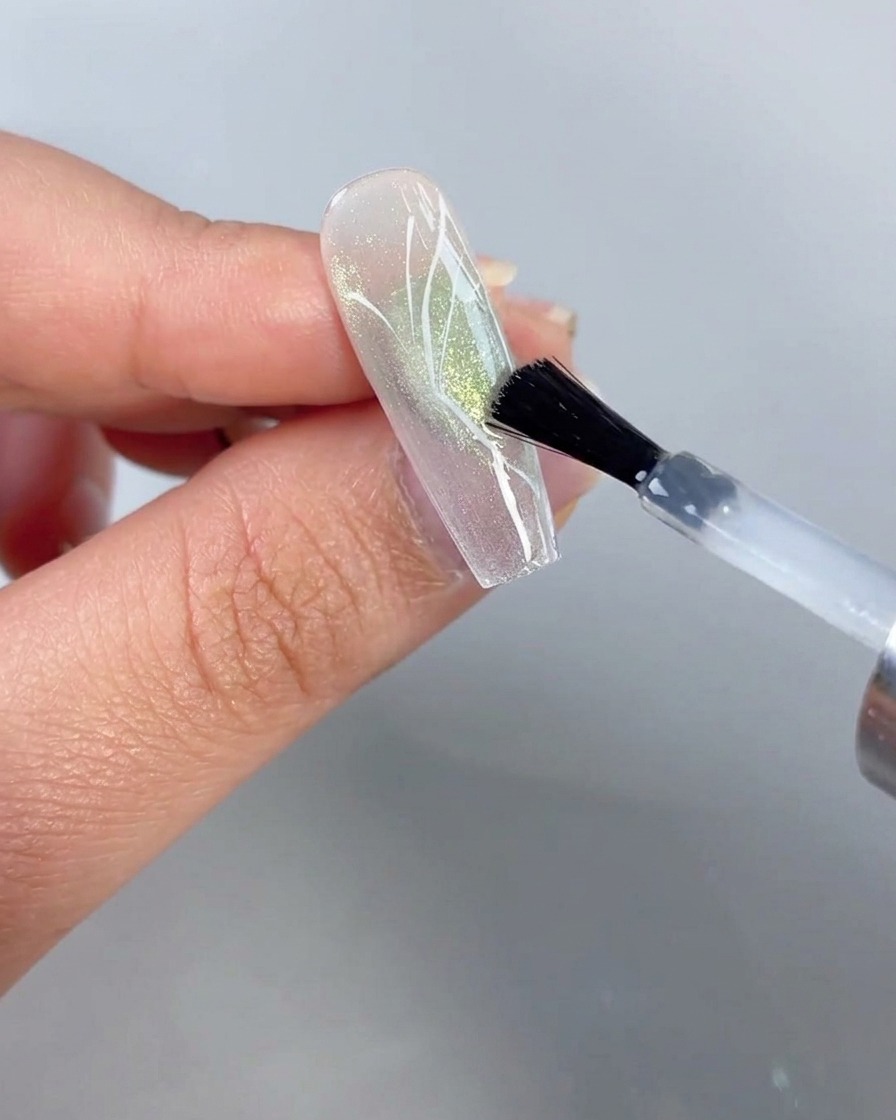 Un pinceau appliquant une couche de finition transparente sur un ongle présentant un design de demi-aile de papillon peint sur fond œil-de-chat vert diaphane.