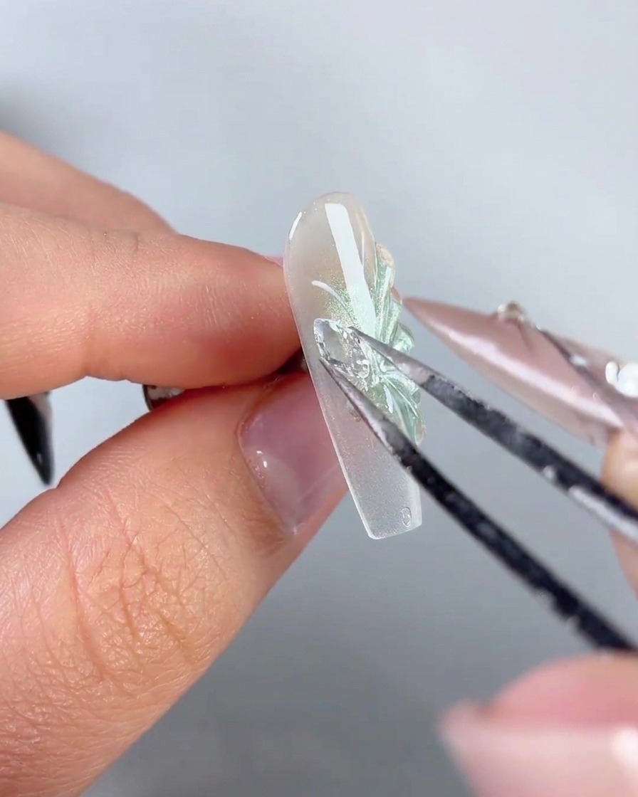Une paire de brucelles métalliques plaçant un charm daile de papillon en strass étincelant sur le côté dun ongle décoré.