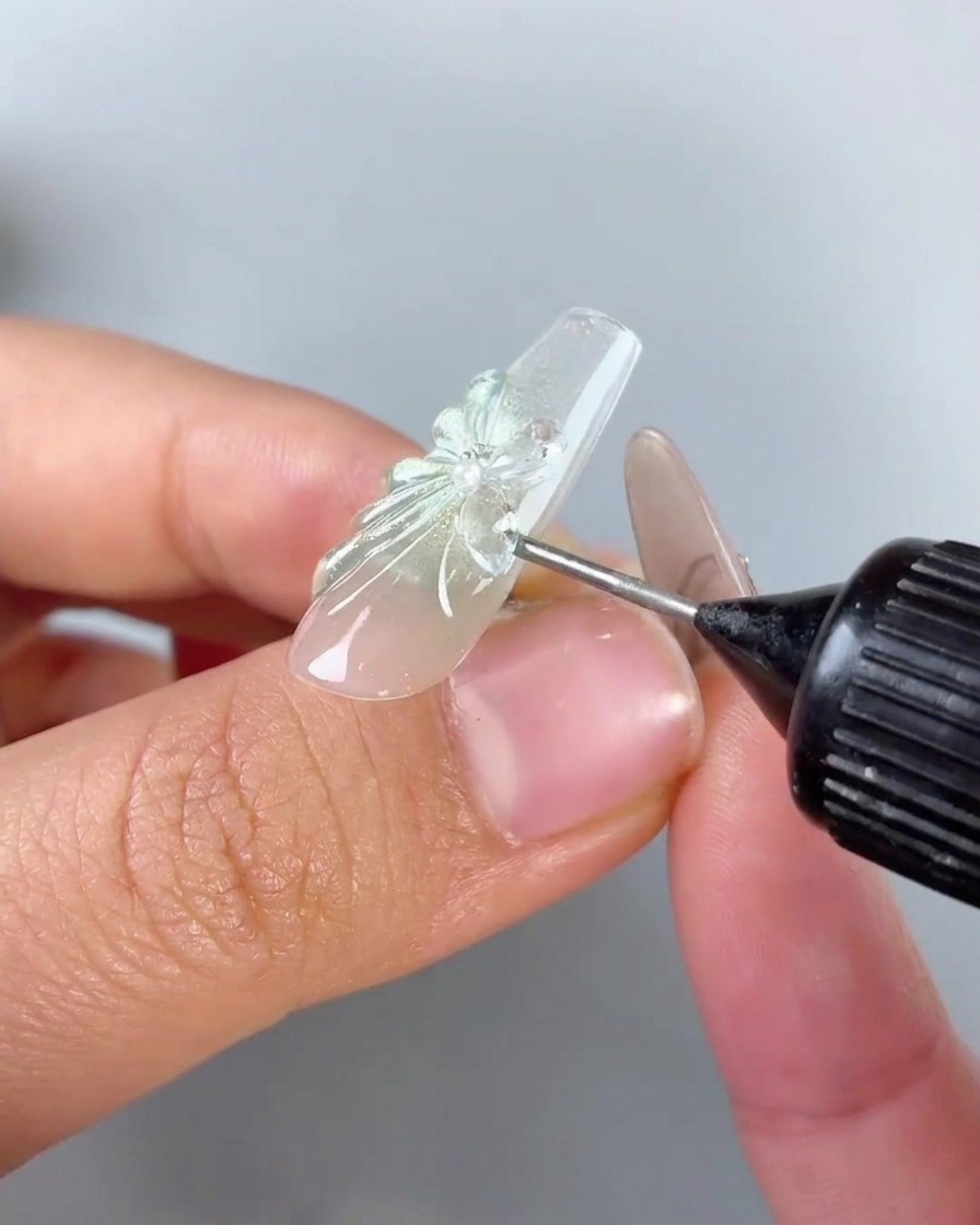Un flacon distributeur à embout de précision appliquant du gel de comblement transparent autour de la base et des bords dun charm de papillon en strass sur un ongle.
