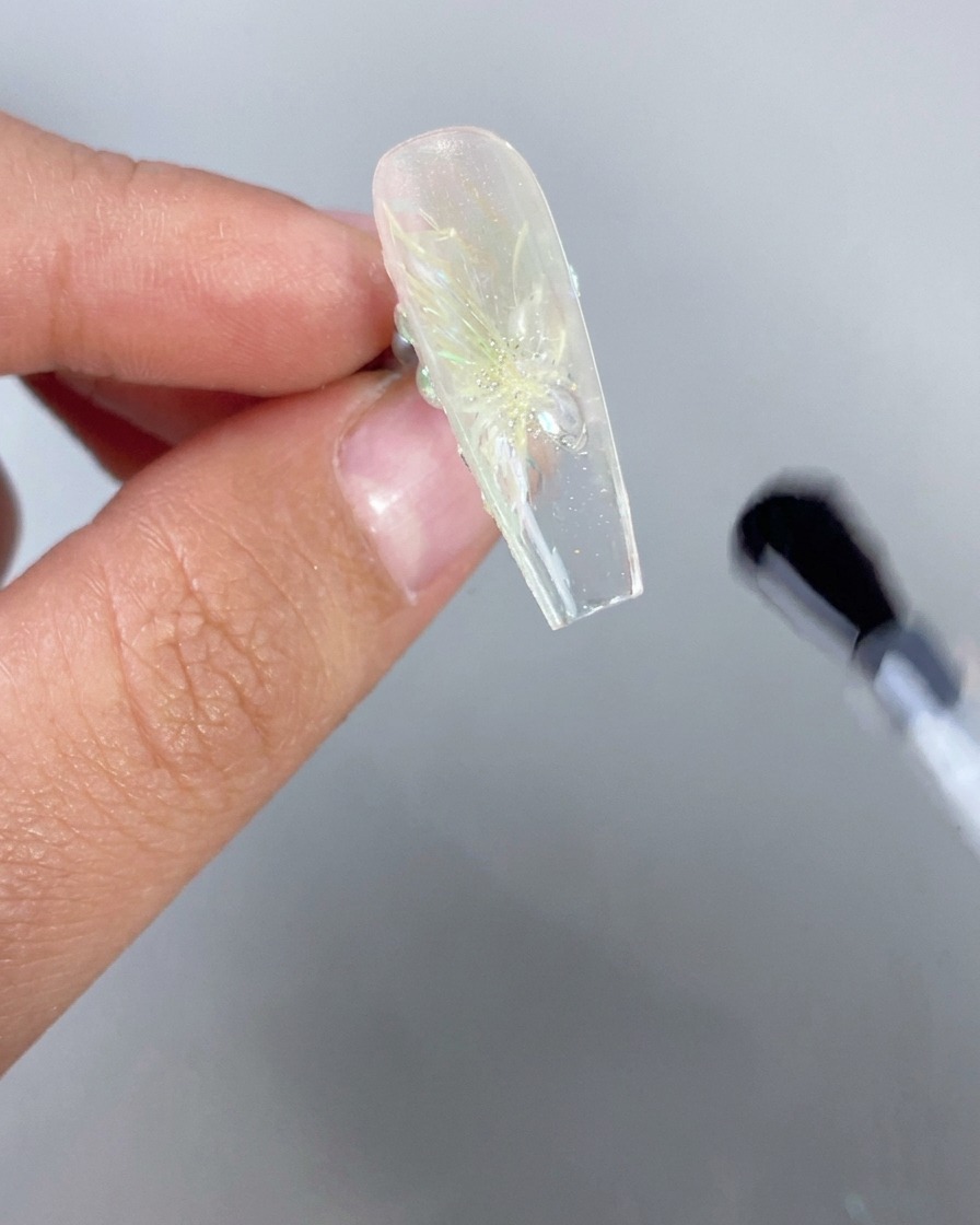 Gros plan dune main tenant à lenvers un ongle transparent effet glacé avec un design floral 3D, tandis quun pinceau applique une finition transparente sur le dessous du bord libre.