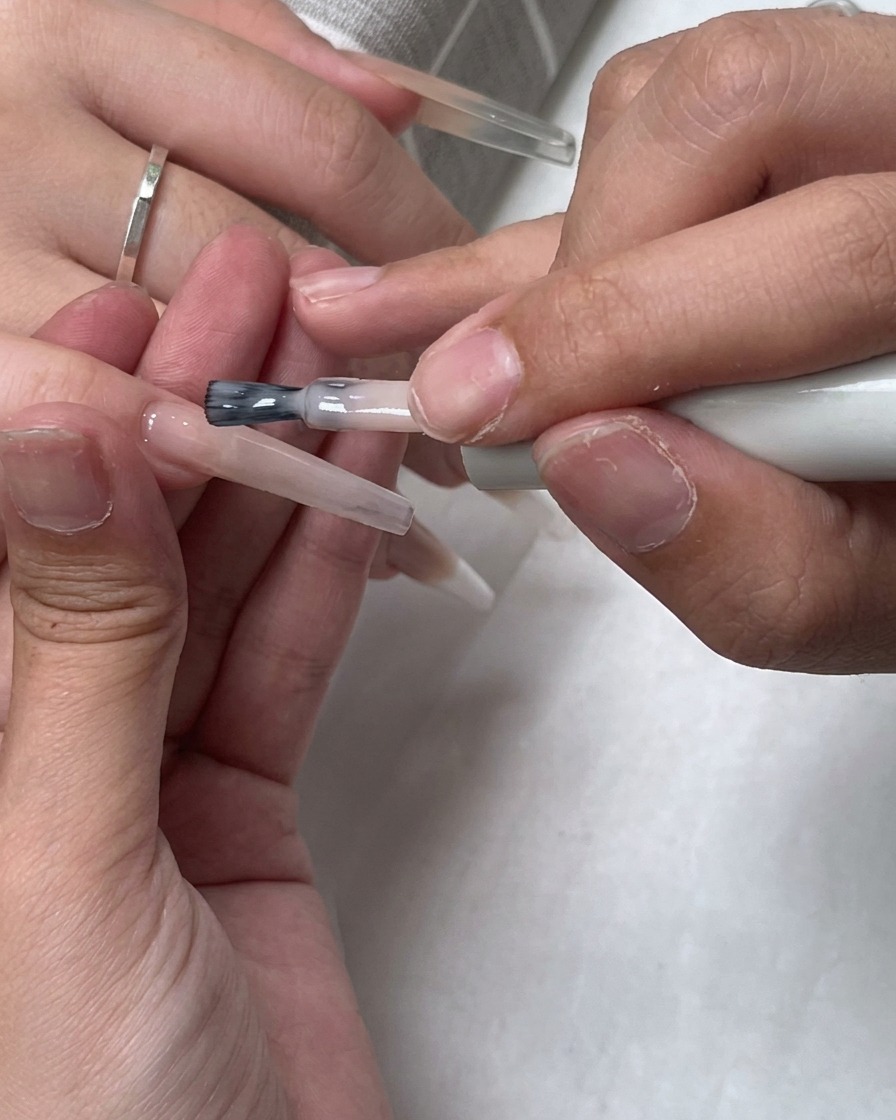 Un pinceau appliquant une couche de vernis nude diaphane sur toute la surface de longle.
