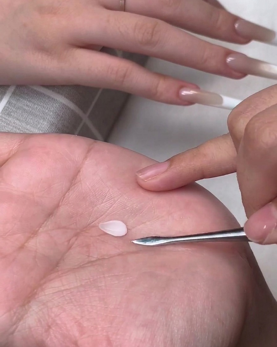 Un petit pétale en gel 3D blanc en forme de goutte posé sur la paume dune personne à côté dun outil de modelage métallique.
