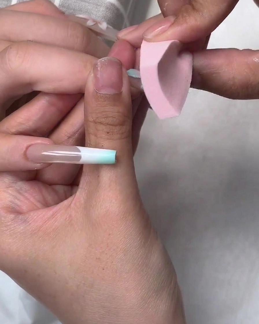 Une petite éponge rose tamponnant du vernis gel bleu sur les bords dune application dongle lors dun tutoriel de nail art.
