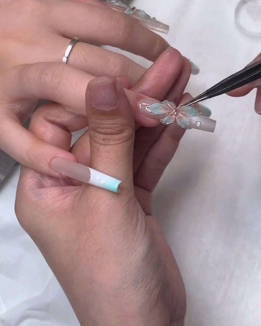 Un gros plan montrant une pince fine plaçant précisément un petit bijou en strass au centre dune fleur en gel sculpté 3D translucide sur un long ongle carré.