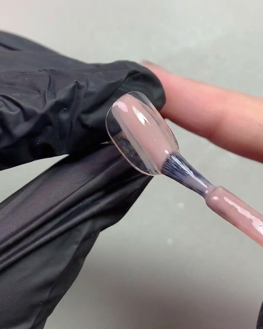 Gros plan dune main gantée utilisant un pinceau pour appliquer un gel nude transparent aux notes orangées sur une capsule dongle.