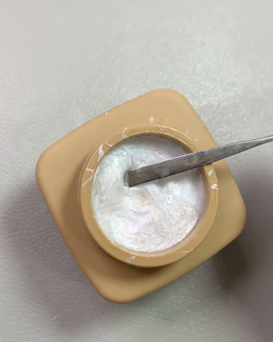 Un outil spatule en métal prélevant du gel de modelage nacré irisé dans un petit pot beige.