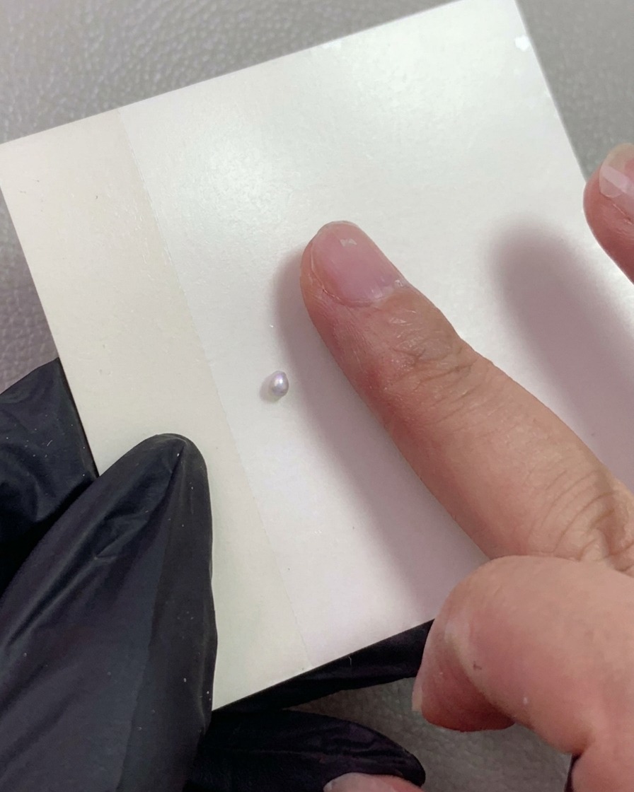 Une main gantée pointant vers une petite boule de gel de modelage nacré posée sur une feuille de mélange.
