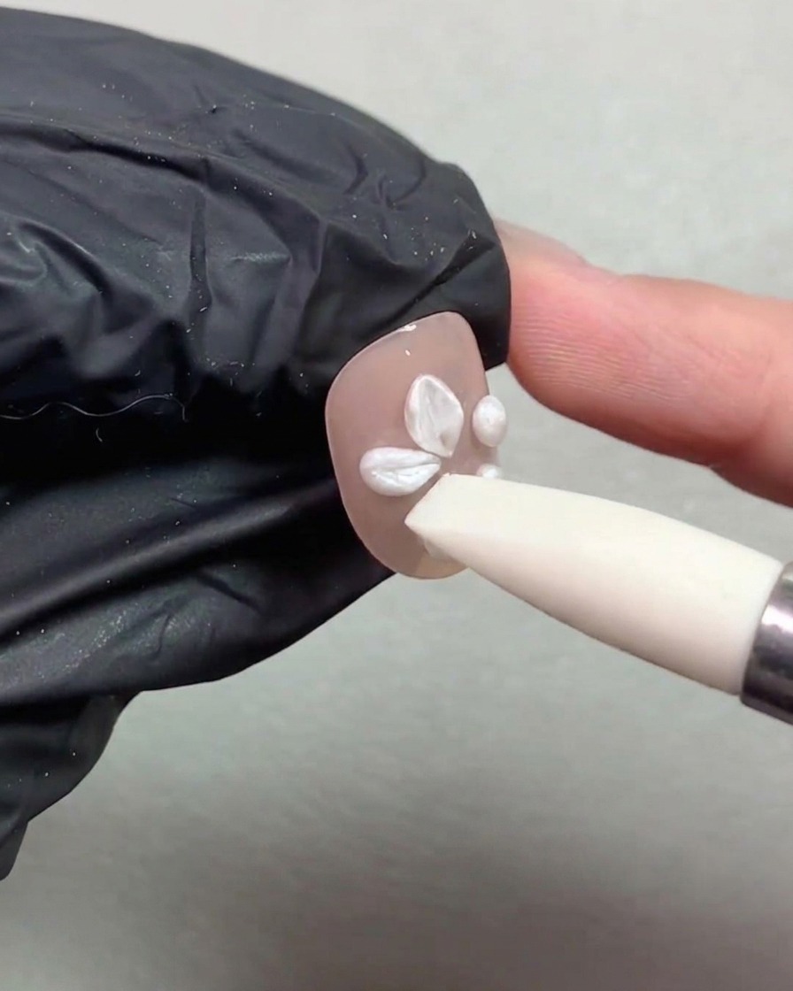 Un outil stylo en silicone pressant et façonnant des boules de gel blanc perlé en pétales de fleurs sur un ongle peint en nude.