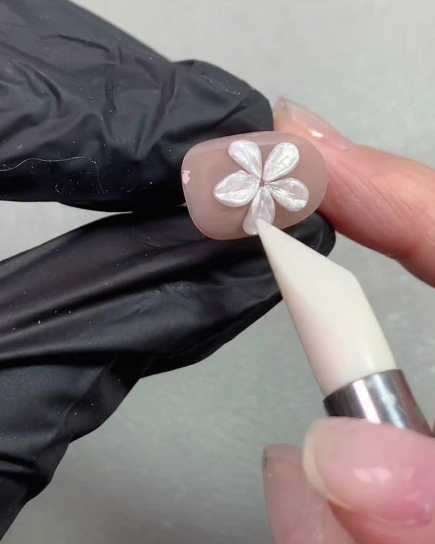 Un stylo de modelage en silicone lissant et ajustant la forme des pétales dune fleur nacrée sur un ongle de couleur nude.