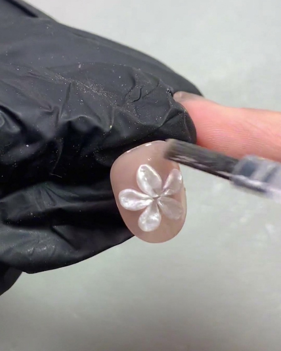 Une main gantée plaçant un ongle court orné dune fleur en perle blanche 3D sous une lampe de polymérisation UV/LED violette.
