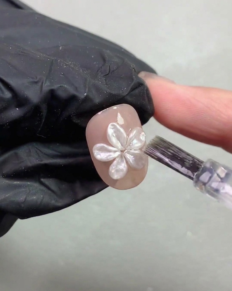 Un pinceau à ongles appliquant délicatement une fine couche de top coat autour dune fleur nacrée 3D catalysée sur un ongle.