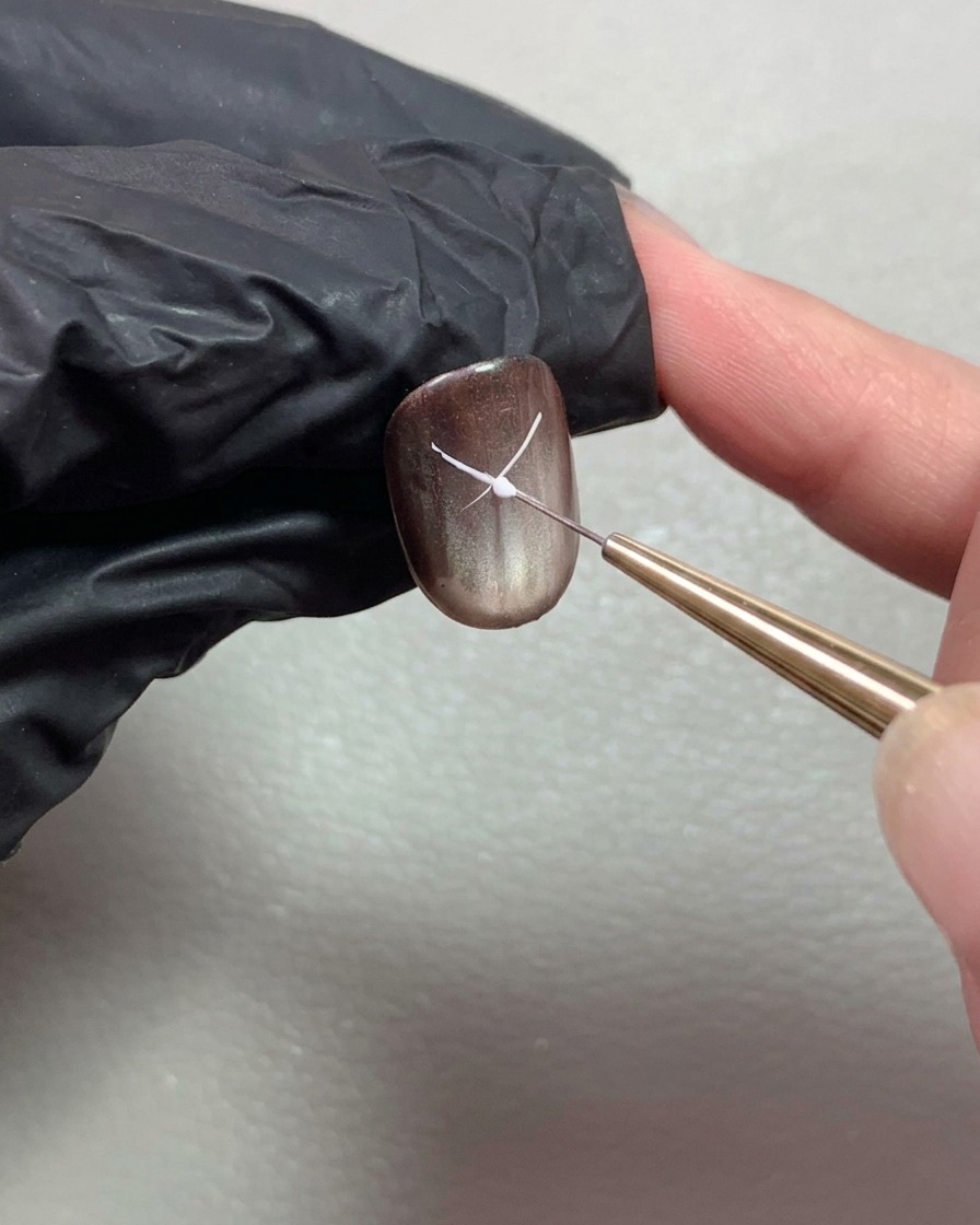 Un pinceau liner fin dessinant de fines lignes blanches pour commencer à former un nœud sur un ongle brillant marron foncé.