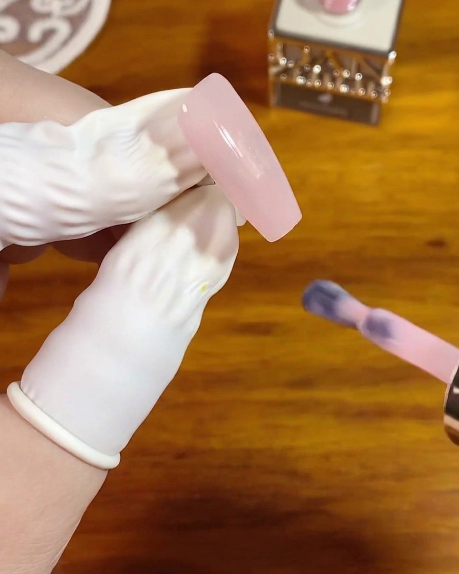 Un pinceau à ongle appliquant une couche lisse de gel cristal rose translucide sur un ongle nu et long de forme carrée.