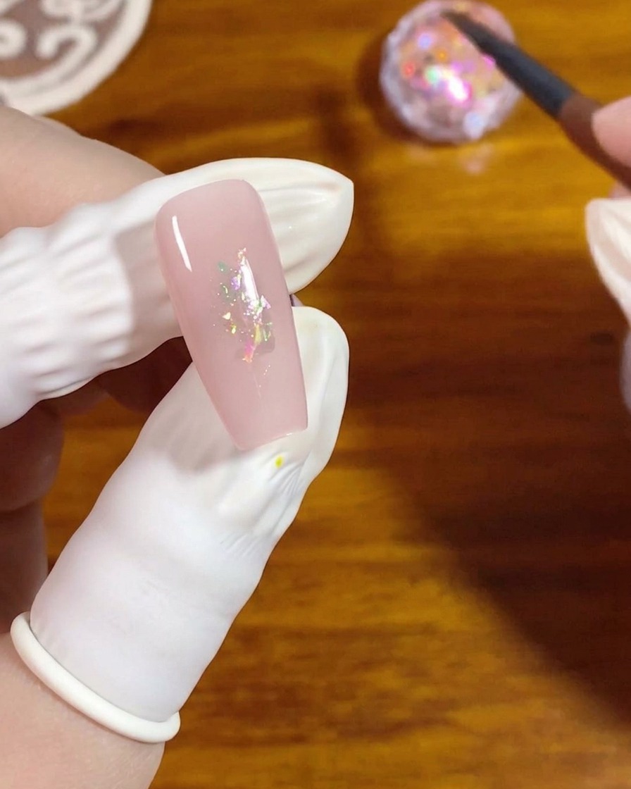 Un pinceau gel plat pressant de petites paillettes opale iridescentes au centre dun ongle vernis en rose.
