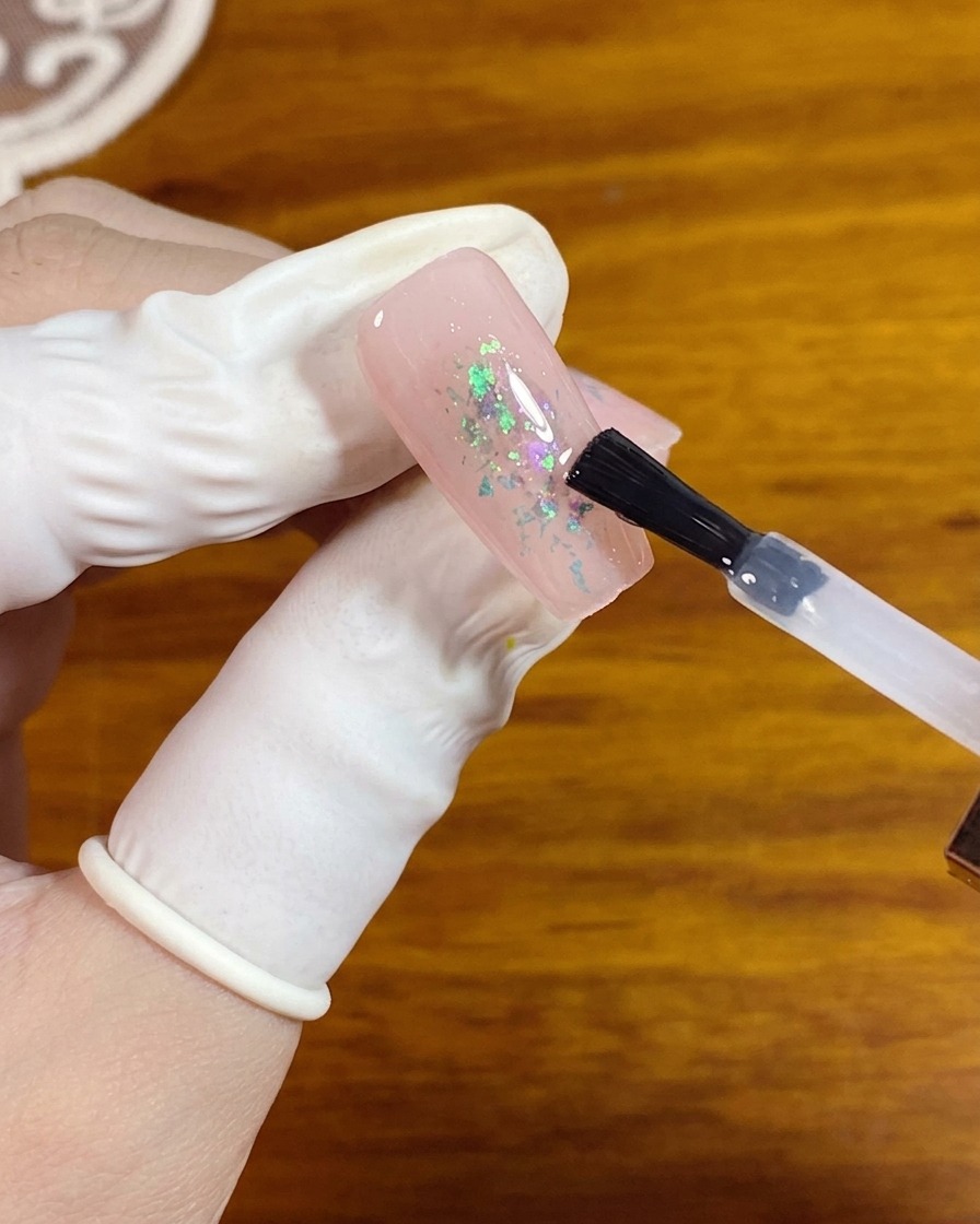 Un pinceau gel appliquant un top coat mat transparent sur un ongle rose avec des paillettes opale au centre.