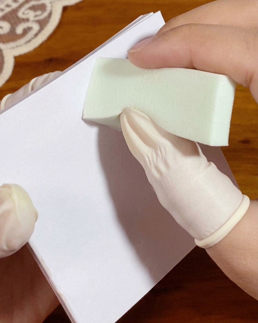 Une main gantée pressant une éponge chargée de gel blanc sur un papier palette avant lapplication sur longle.
