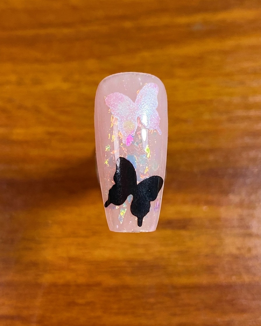 Vue de haut dun ongle décoré avec des paillettes iridescentes et deux formes de pochoirs papillons.