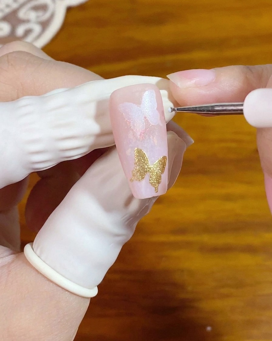 Un pinceau de détail fin appliquant de minuscules points blancs à côté dun élément de nail art papillon doré.