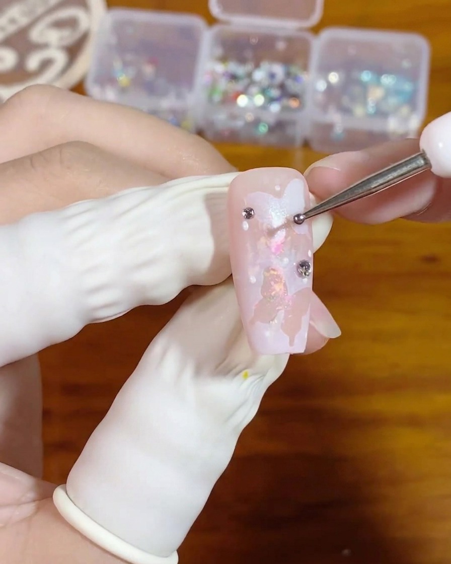 Gros plan dune main utilisant un outil de dotting fin pour placer une minuscule perle de gel transparent sur un ongle rose décoré dun art de papillon blanc.