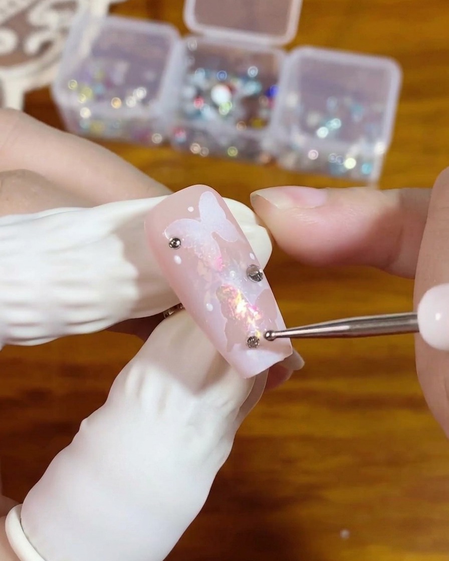 Un outil de dotting plaquant un petit strass argenté à dos plat sur une perle de gel transparent sur un ongle rose présentant un design de papillon.