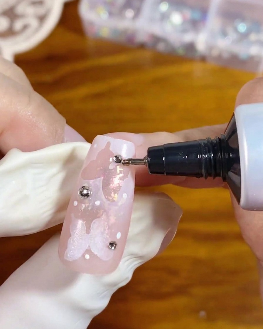 Un tube applicateur à embout de précision distribuant du gel transparent autour de la base dun strass argenté sur un ongle rose décoré pour le fixer en place.