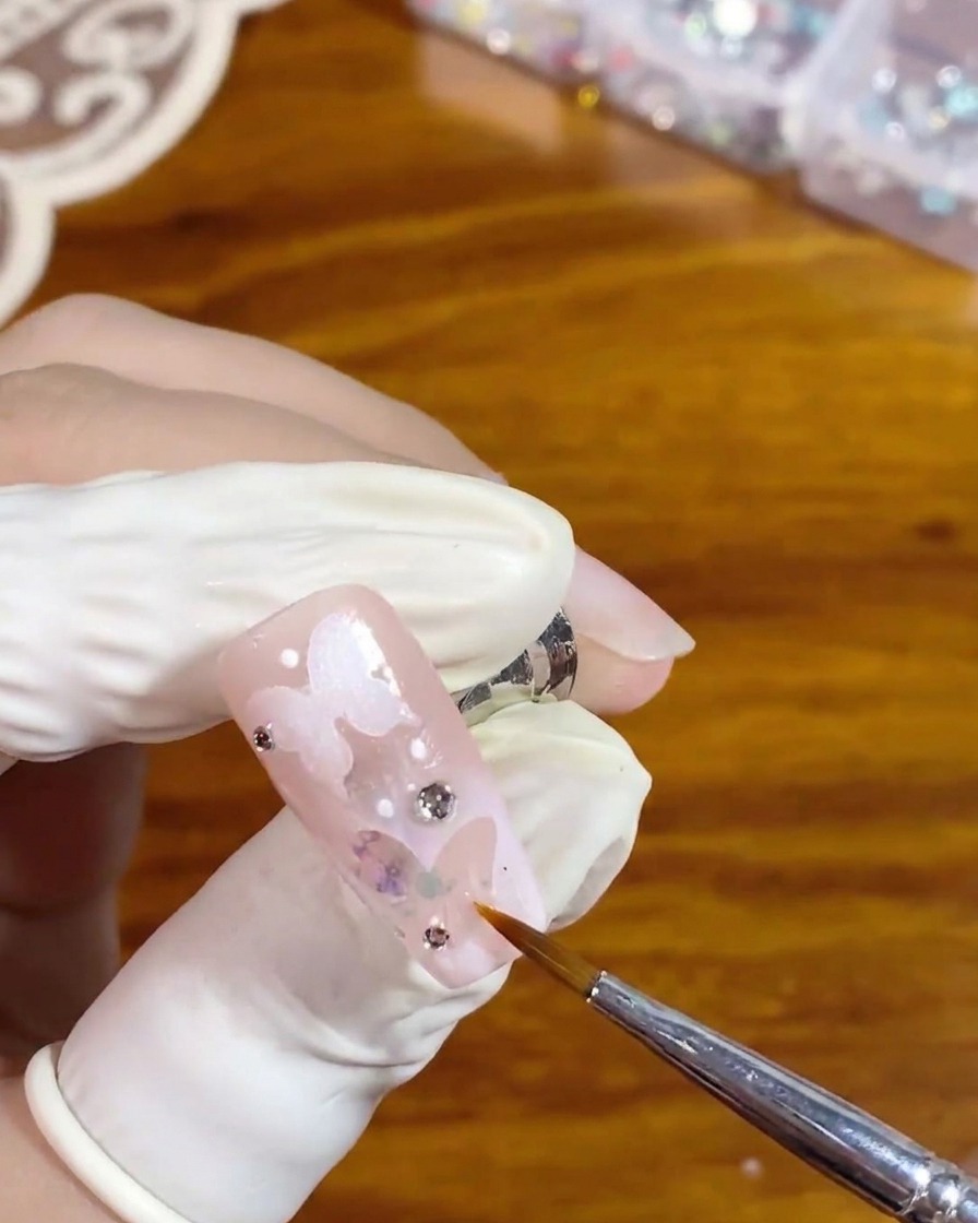 Un pinceau liner fin appliquant soigneusement un top coat brillant transparent autour dun strass argenté sur un ongle rose avec un design de papillon évidé blanc.