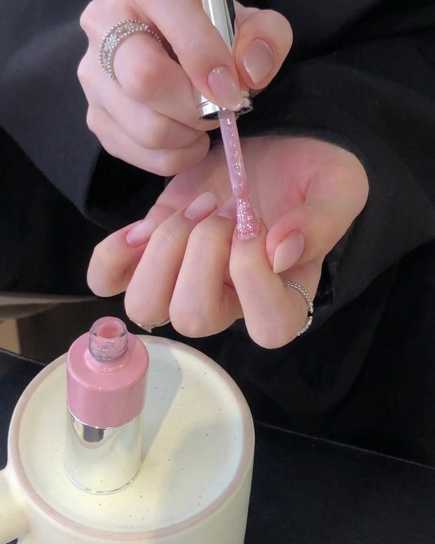 Paire de mains tenant un pinceau de vernis rose au-dessus dongles manucurés avec un flacon rose reposant sur un support en céramique.
