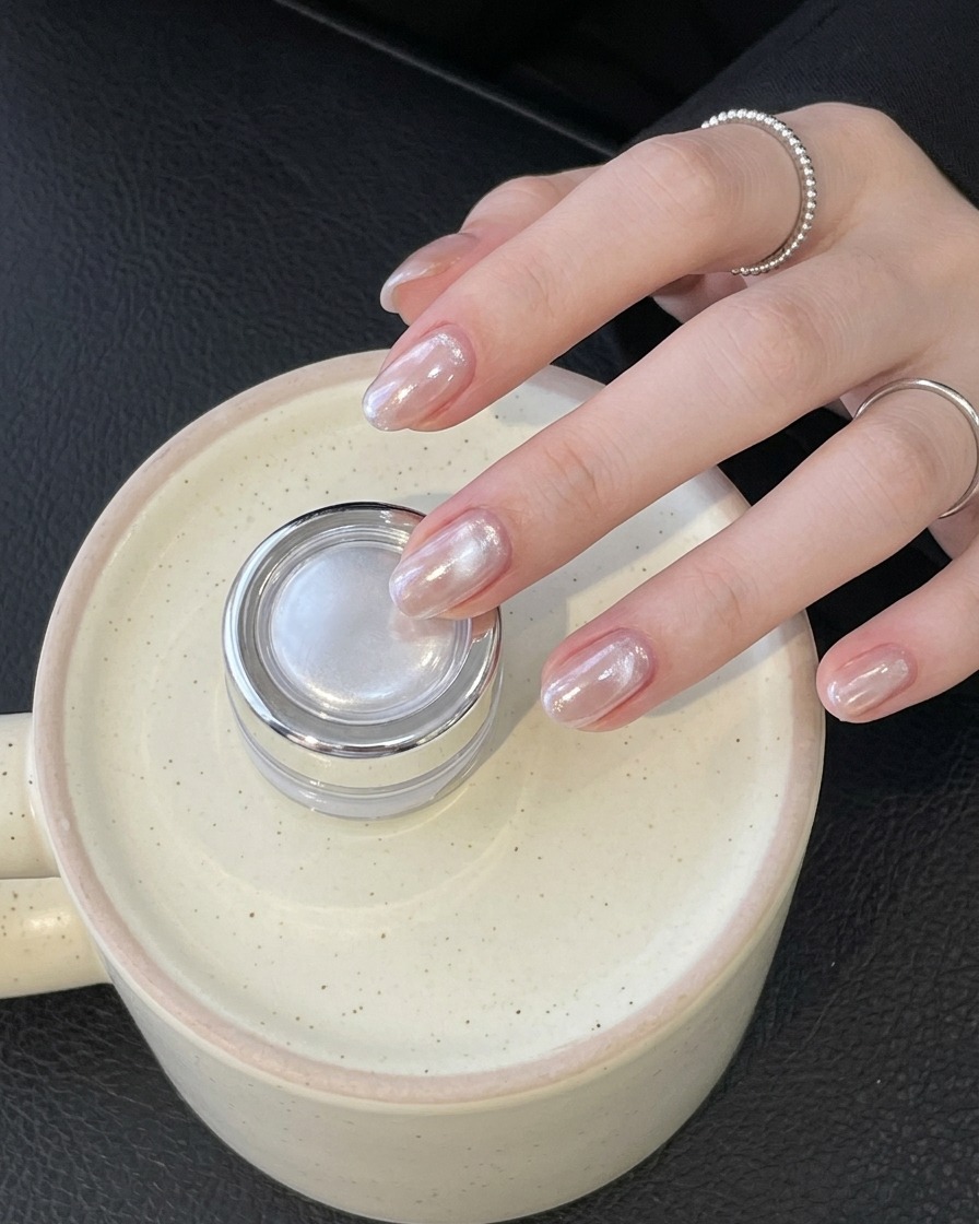 Main portant une bague tenant un petit pot argenté tout en présentant des ongles chromés brillants finis.