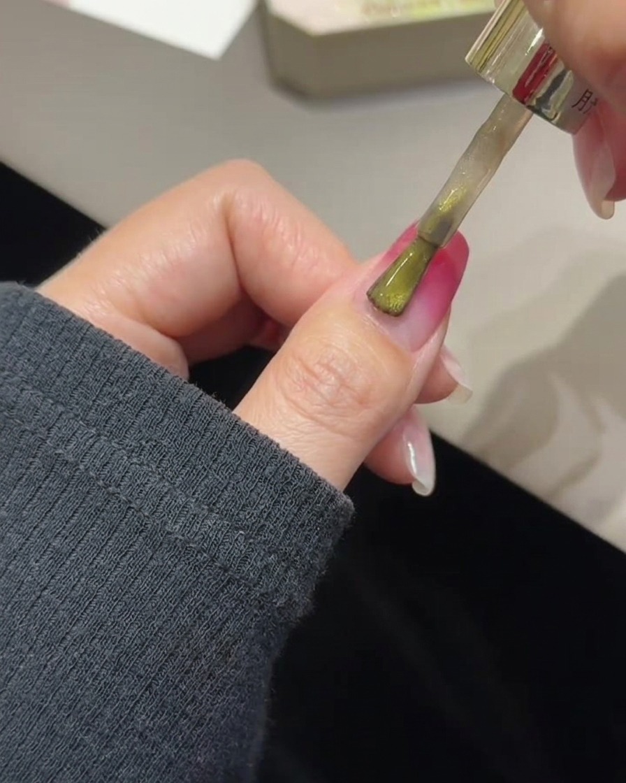 Un pinceau appliquant du gel œil de chat doré sur un ongle avec dégradé rose déjà catalysé.