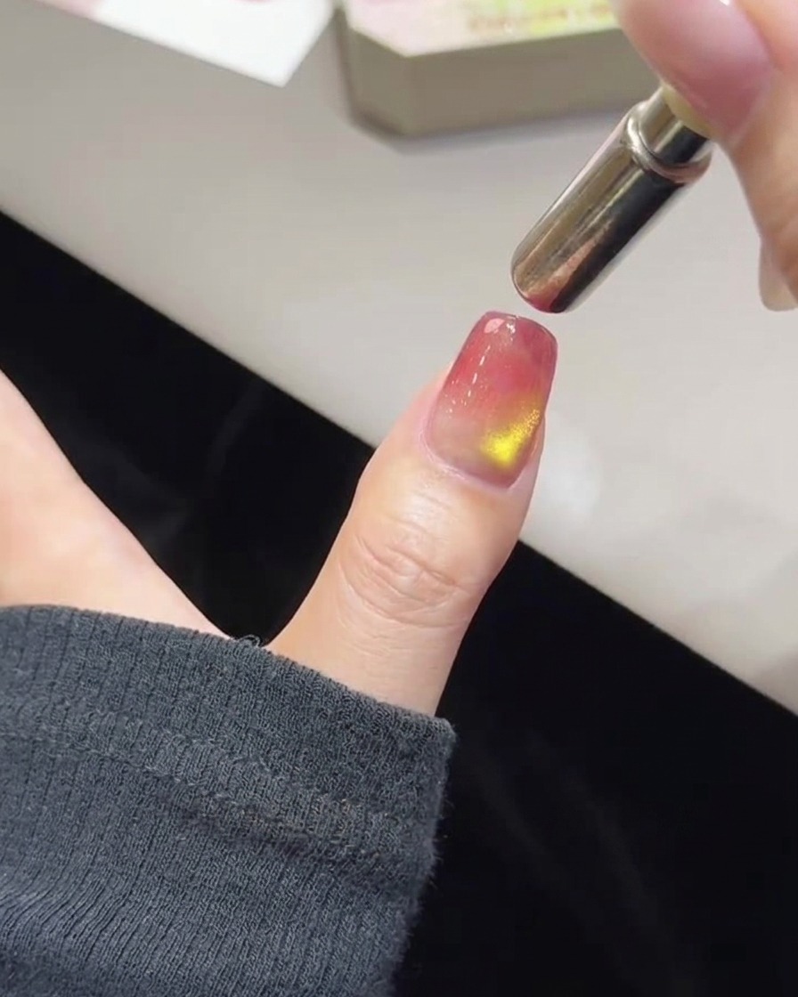 Gros plan dun pouce avec du vernis rose et doré non catalysé et une baguette magnétique cylindrique maintenue au-dessus du bord de longle.