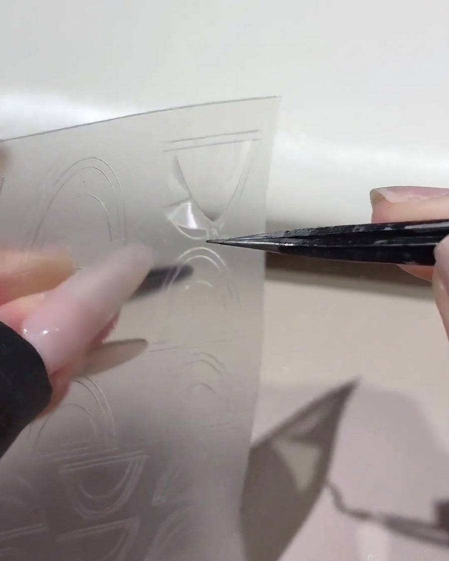 Une paire de pinces soulevant un autocollant guide French incurvé dune feuille de plastique transparent.