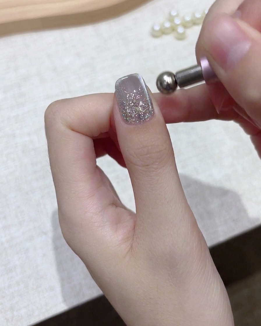 Gros plan dune main tenant un outil magnétique sphérique au-dessus dun ongle en gel à paillettes argentées.