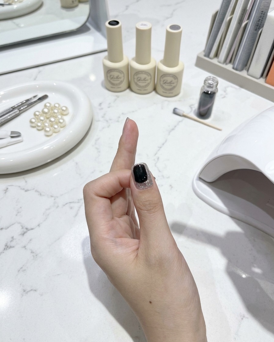 Un pinceau à ongles appliquant une couche de finition brillante sur un ongle œil-de-chat fini avec des outils de manucure et des flacons de vernis sur une table en marbre blanc en arrière-plan.